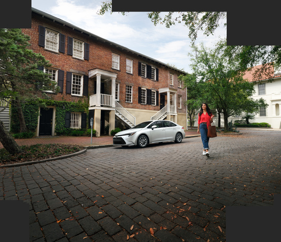 A woman in a red blouse and jeans walks away from a white Toyota Corolla parked in front of a residence.