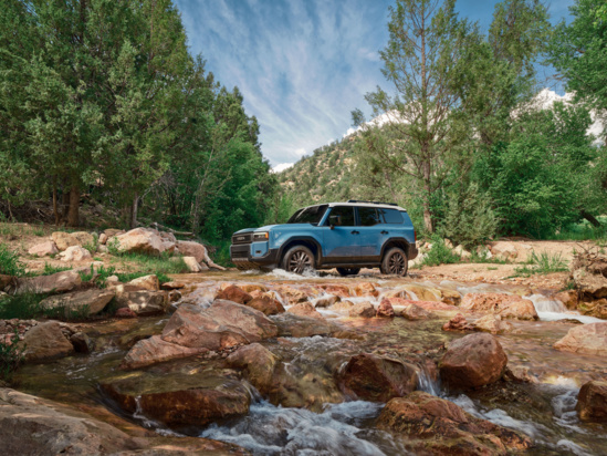 A blue Toyota Land Cruiser is parked near a river in the woods.